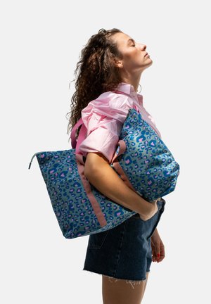 Woman with curly hair wearing a pink shirt and denim skirt holding blue and pink leopard print tote bags against a plain background.