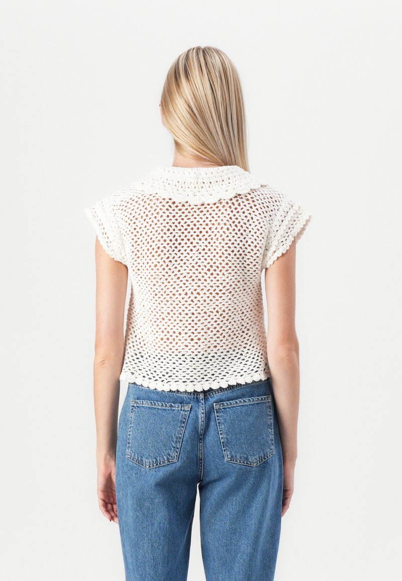 Weißes, offen gestricktes kurzärmeliges Oberteil mit gewelltem Kragen und Saum, kombiniert mit hochgeschnittenen blauen Jeans.