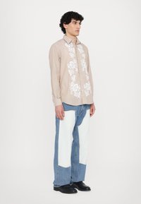 Camicia beige con bottoni e ricami floreali bianchi, abbinata a jeans blu con pannelli bianchi. Il modello è in piedi rivolto a sinistra.