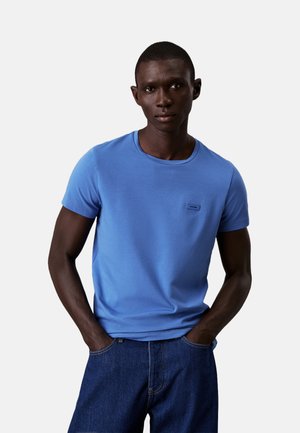 Camiseta de algodón azul con cuello redondo, mangas cortas y un pequeño parche de logo en el pecho. Unos jeans de mezclilla oscura completan el conjunto.
