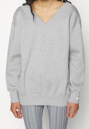 Femme portant un sweat-shirt gris clair à col en V avec poignets et ourlet côtelés, associé à un pantalon gris rayé, debout sur fond blanc.