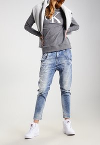 Haut à capuche gris avec logo, superposé à une chemise grise, associé à un jogging en denim délavé et des baskets blanches. Style décontracté, coupe ample.