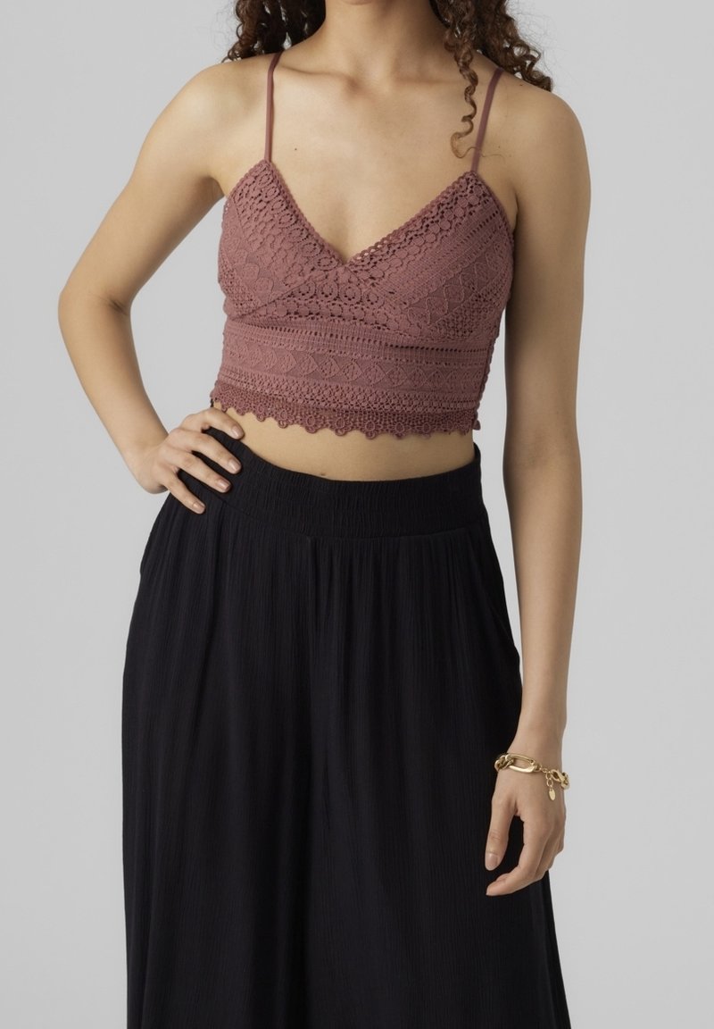 Kvinna klädd i en mauve virkad crop top med tunna axelremmar och högmidjade vida svarta byxor, stående med en hand på höften.