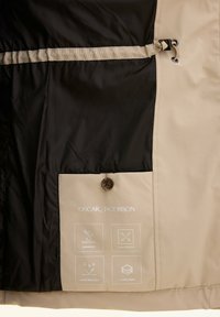 Beige en zwart jakkeren interieur met winddichte label. Bevat een met knopen afsluitbare zak, iconen voor waterafstotend, 4-way stretch en 3-laags stof.