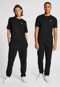 Svarta bomulls-t-shirts visas med korta ärmar och rund hals, kombinerade med svarta mjukisbyxor, båda med små guldiga logotypdetaljer.