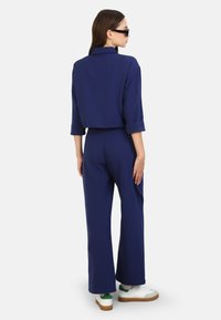 Marinblus med kort ärm och uppvikta ärmar, tillsammans med matchande vida byxor. Modellen har vita sneakers med gröna detaljer och svarta solglasögon.