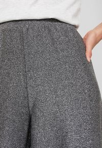 Hoge taille, grijze glinsterende broek met een elastische tailleband en zijzakken, met een gestructureerde stof. Close-up van het taillegebied.