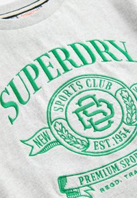 Grå sweatshirt med iögonfallande gröna broderade bokstäver och ett runt emblem med detaljer. Ribbad halsringning, avslappnad sportklubbsdesign.
