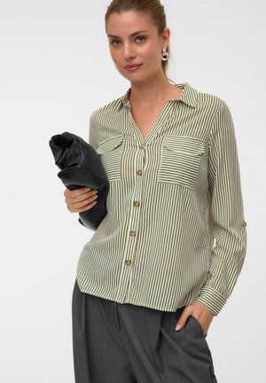 Frau trägt ein grün-weiß gestreiftes Hemd mit Knöpfen und graue Hose und hält eine schwarze Clutch vor weißem Hintergrund.