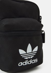 Zaino in tessuto nero con superficie strutturata, caratterizzato da un logo Adidas bianco e tre strisce orizzontali. Chiusura con zip nella parte superiore.