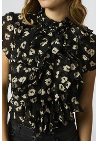 Femme portant un chemisier noir transparent avec des motifs floraux blancs et des volants superposés à l'avant et des manches courtes.