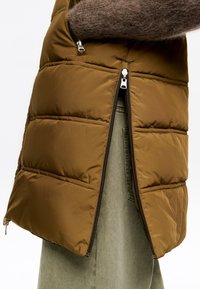 Chaleco acolchado marrón con cremallera lateral parcialmente abierta, mostrando pantalones verde oliva y la manga de un suéter marrón de felpa.