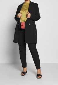 Svart lång kavaj över en senapsgul topp, ihop med svarta byxor och svarta sandaler. En liten röd skulderväska bärs vid midjan.