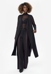 Kobieta o kręconych włosach, ubrana w przezroczystą czarną bluzkę, długi czarny bezrękawnik, szerokie czarne spodnie z ciemnymi pionowymi paskami oraz czarne buty na szpic, stojąca.