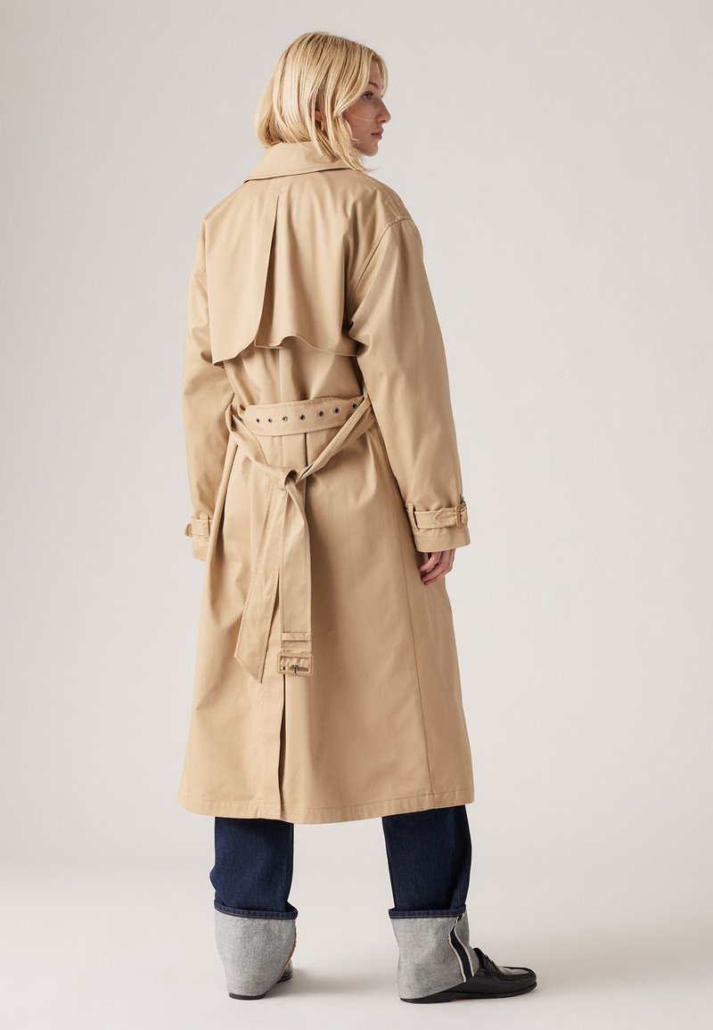 Beige Trenchcoat mit Rückenklappe, tailliertem Gürtel und hochrollbaren Ärmeln, kombiniert mit dunklen Jeans und grauen Knöchelstiefeln. Glatte Stofftextur.