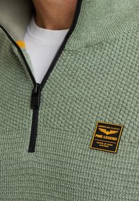 Groene pullover met een gestructureerd breiselontwerp, zwarte rits en een geel en zwart logo-opdruk op de borst. Heeft een hoge kraag.