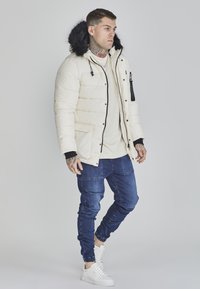 Creme puffer-jakke med hætte i sort pels, lynlåse foran og elastiske mansjetter. Bæres med mørke denim joggers og hvide sneakers.