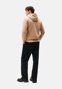 Joven de pie con las manos en los bolsillos, vistiendo una sudadera beige, pantalones oscuros y zapatos negros, mirando ligeramente hacia la izquierda sobre un fondo blanco.