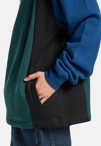 Sweatshirt em blocos de cor com um design em verde, azul e preto. Material de algodão macio com um bolso frontal e detalhes de costura em contraste.