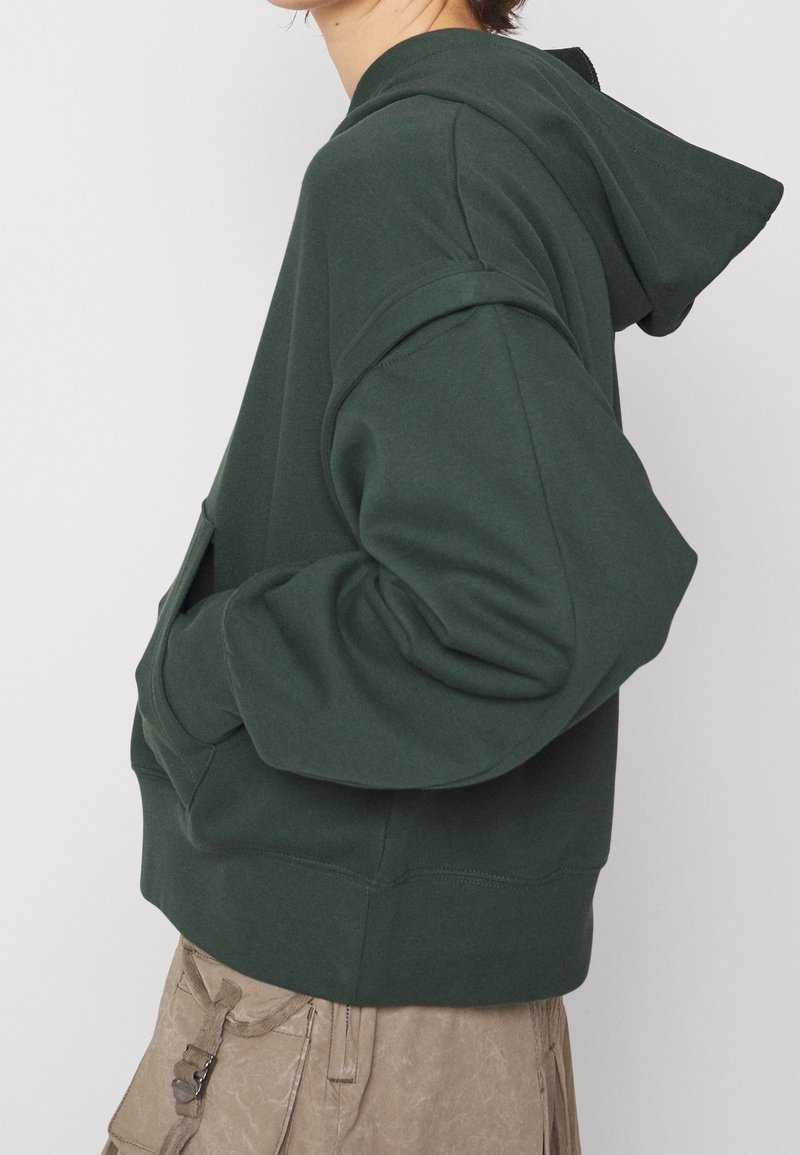 Personne portant un sweat à capuche oversize vert foncé avec les mains dans la poche avant et un pantalon cargo beige, vue de profil.