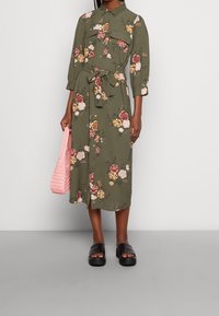 Grünes florales Midikleid mit durchgehendem Knopfverschluss, Bindegürtel und zwei Brusttaschen; gestylt mit einer gewebten rosa Handtasche und schwarzen Sandalen.