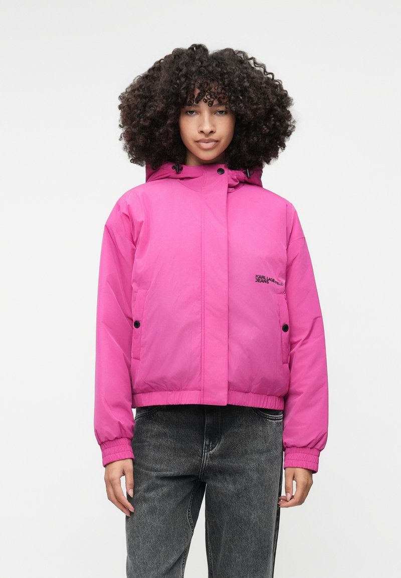 Jachetă izolată roz cu glugă, dotată cu manșete elastice, un design cropped și un detaliu cu logo pe partea stângă a pieptului. Purtată peste blugi gri închis.