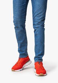 Scarpe da atletica rosse con una parte superiore texturizzata, cordini e una suola intermedia bianca, indossate con jeans blu attillati.