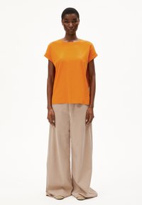 T-shirt orange à manches courtes en tissu doux, associé à un pantalon large beige. Le T-shirt présente un détail de logo subtil.