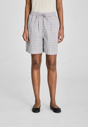 Osoba oblečená v béžovom tielku bez rukávov, bielo-sivých kockovaných kraťasoch po kolená so sťahovacou šnúrkou a čiernych balerínach, stojaca pred jednoduchým pozadím.