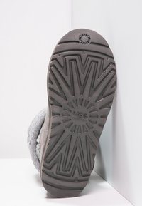 Suela de una bota UGG gris con un dibujo en la suela, mostrando el nombre de la marca y una parte superior de tela de punto contra un fondo blanco.