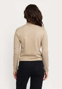 Beige, langärmliger Pullover mit geripptem Kragen und körperbetontem Schnitt. Getragen mit schwarzen Jeans, die eine glatte Textur und neutrale Farbe zeigen.