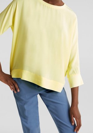 Persoon draagt een loszittende lichtgele top met lange mouwen en brede manchetten en blauwe aansluitende broek, hand op de heup, tegen een lichte achtergrond.