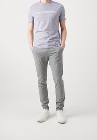 T-shirt con collo rotondo di colore viola chiaro con logo bianco, abbinata a pantaloni slim-fit grigi chiari e sneakers bianche, su uno sfondo neutro.