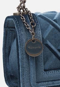 Blå sammetsväska med kviltad textur, med en kedjesladd och en rund bricka med varumärket "Tamaris."