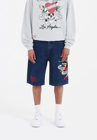 Shorts de denim en azul oscuro con diseños bordados de un pájaro y pancartas. Combinados con una sudadera gris con gráficos coloridos. Zapatillas blancas.