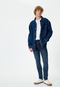 Camisa de mezclilla en azul oscuro, con botones y un bolsillo en el pecho, combinada con jeans oscuros ajustados y zapatillas blancas con suela beige.
