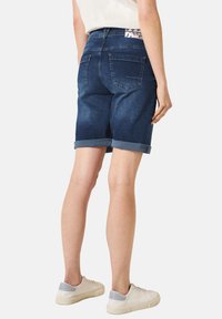 Shorts en denim bleu marine avec ourlets roulés, cinq poches et une ceinture à motif blanc. Associés à des baskets de couleur claire.