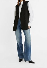 Schwarzes doppelt geknöpftes Weste mit Schulterpolstern über einem weißen Hemd, kombiniert mit hellblauen weit geschnittenen Jeans und schwarzen spitzen High Heels.