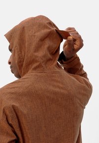 Giubbotto con cappuccio di tessuto marrone a trama, con coulisse. Il tessuto presenta un motivo sottile. Il modello è visto mentre aggiusta il cappuccio da dietro.