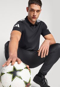 Svart polo skjorte med hvite striper på skuldrene, svarte treningbukser, holder en fotball med stjernedesign, og svarte fotballsko.