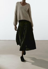 Pull beige en tricot avec col en V et manches tombantes, associé à une jupe plissée noire. Chaussures noires à enfiler avec des accents cloutés.