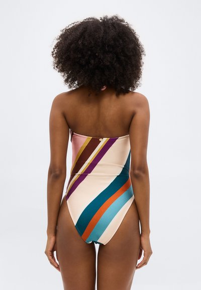 Γυναίκα με σγουρά μαλλιά, που φοράει ολόσωμο μαγιό με πολύχρωμες ρίγες χωρίς τιράντες, γυρισμένη από την πλάτη, μπροστά σε απλό φωτεινό φόντο.