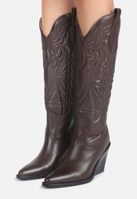 Stivali da cowboy in pelle marrone con punta affilata, motivi di cucitura intricati e zeppa; caratterizzati da altezze elevate e tappi ripiegati.