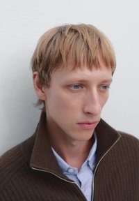 Brązowy sweter z ribbowanym splotem i wysokim kołnierzem, noszony na jasnoniebieskiej koszuli. Modelka ma krótkie blond włosy i jasnoniebieskie oczy.