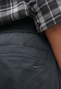 Camisa de flanela xadrez preto e cinza cobrindo parcialmente calças cinza escuro com textura suave, apresentando um bolso traseiro e um pequeno logótipo da Nike.