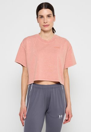 Femme aux cheveux foncés attachés, portant un t-shirt court rose délavé et un pantalon de survêtement gris Under Armour avec des bandes blanches sur les côtés.