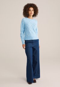 Lichtblauwe gebreide trui met een golfpatroon, gecombineerd met donker brede jeans. De outfit heeft een ontspannen pasvorm en lange mouwen.