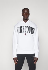 Sudadera blanca de Nike con "NIKE COURT" en gran tamaño y en negro. Cuenta con capucha ajustable y un corte relajado. Combinada con pantalones negros con detalles del logo.