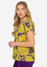 Top de manga corta en un vibrante color amarillo con patrones florales morados. Presenta un escote en V y un corte relajado, fabricado en una tela ligera con una textura suave.