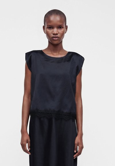 Femme aux cheveux courts portant un haut en satin noir sans manches avec bordure en dentelle et une jupe noire assortie, sur un fond blanc uni.